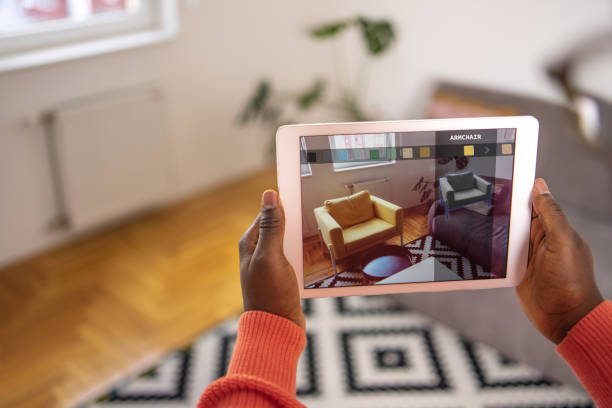 Man's hands hold a digital tablet with the augmented reality app, which shows the armchair in the living room. In the app, you can choose the model of a chair and the color and texture of the fabric.